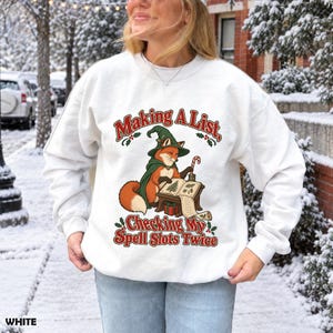 Puede incluir: Una sudadera blanca de manga larga con el gráfico de un zorro con sombrero y capa de mago verde, escribiendo en un libro. El texto "Making A List, Checking My Spell Slots Twice" aparece en letras rojas decorado con hojas de acebo.