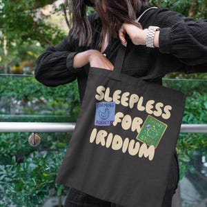 May include: Black tote bag with the text "Sleepless for Iridium". The bag has two patches: one with a chicken and the words "Farming Addict", and another with a green creature and "Shiny Stuff". The bag is held by a person.