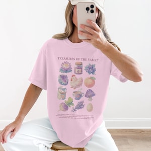 Könnte beinhalten: Hellrosa T-Shirt mit dem Text "Treasures of the Valley" und verschiedenen Illustrationen von Lebensmitteln und Pflanzen. Die Illustrationen umfassen ein Glas Marmelade, ein Huhn und einen Pilz. Das Shirt hat eine lockere Passform.