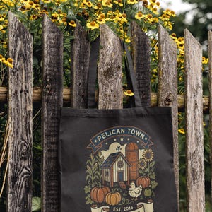 May include: Black tote bag featuring a farm scene with a barn, pumpkins, and a chicken. The text reads "PELICAN TOWN" and "FARM LIFE". The bag is displayed on a weathered wooden fence with yellow flowers in the background.