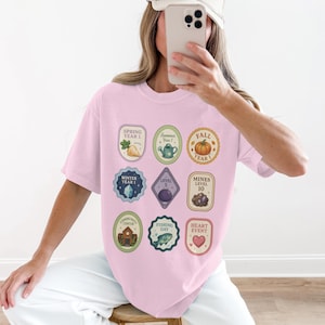Könnte beinhalten: Ein hellrosa T-Shirt mit verschiedenen bunten Abzeichen-Designs. Die Abzeichen enthalten Text und Illustrationen zu Jahreszeiten, Ereignissen und Levels. Das Shirt hat eine lockere Passform und kurze Ärmel.