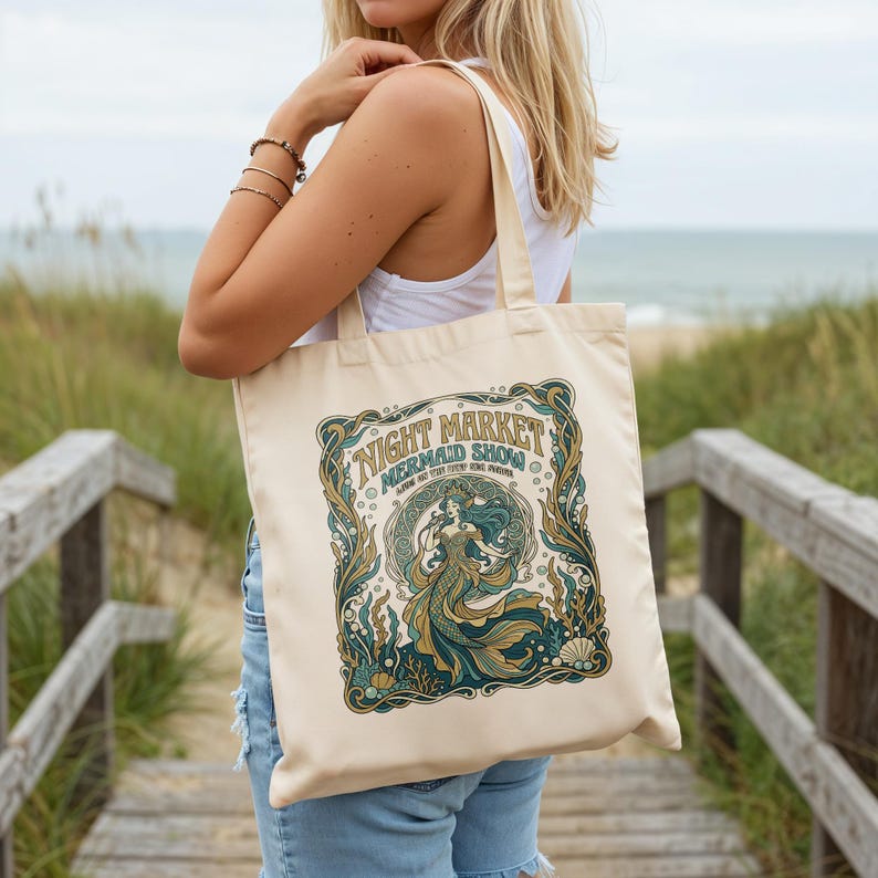 Cream tote bag with mermaid "Night Market Mermaid Show" design on beach boardwalk.