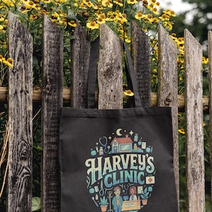 May include: Black tote bag featuring the words "Harvey's Clinic" in a decorative font, with medical-themed illustrations. The bag is suspended on a weathered wooden fence, with yellow flowers visible in the background.
