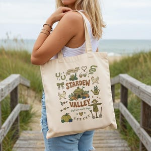 May include: Cream-colored tote bag featuring a colorful illustration with the text "Stardew Valley" and various farm-themed icons. The design includes a scarecrow, chickens, pumpkins, and a watering can. The phrase "Just one more day" is also included.