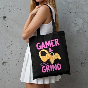 以下が含まれることがあります： 黒いトートバッグ。ピンク色の文字で「GAMER & GRIND」、金色のヘッドセットとゲームコントローラー、そして「all night, every night」の文字が描かれています。バッグには黒いハンドルが付いています。