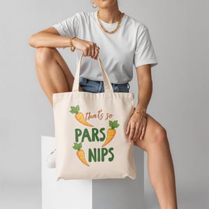 May include: Cream tote bag featuring the text "that's so PARS NIPS" and carrot illustrations. The bag has two handles and is held by a person. The design includes orange carrots with green tops. The bag is made of a durable fabric.