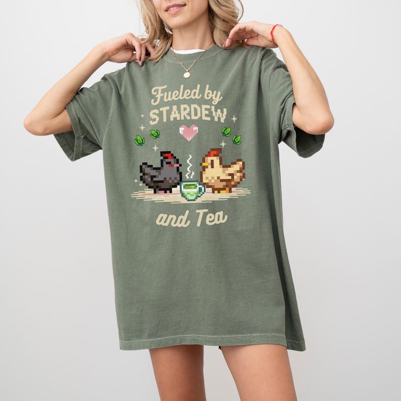 A woman wearing a green t-shirt with a pixelated design of two chickens and a cup of tea, along with the text "Fuelled by Stardew and Tea".