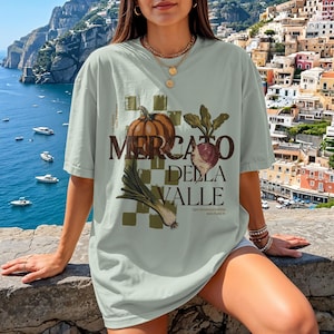 Könnte beinhalten: Hellgrünes T-Shirt mit dem Text "Mercato Della Valle" und Illustrationen eines Kürbis, einer Rübe und eines Lauchs. Das Design befindet sich auf einem karierten Hintergrund. Das Shirt hat eine lockere Passform.