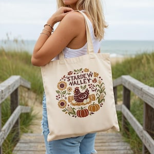 May include: Cream-colored tote bag featuring a circular design with the text "STARDEW VALLEY". The design includes a brown chicken, sunflowers, pumpkins, and leaves. The bag has a simple, rectangular shape with two handles.