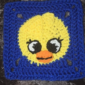 May include: A crocheted square with a yellow duck face, black and white eyes, and an orange beak. The duck is set against a blue background, highlighting a handmade craft item.