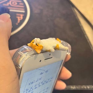 Puede incluir: Una figura de gato blanco y naranja descansando sobre una funda transparente para teléfono. El gato está acostado con los ojos cerrados. El teléfono es blanco y muestra notas manuscritas.