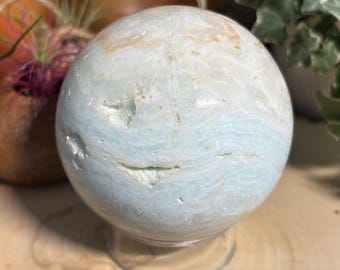 May include: A light blue and white sphere with a marbled appearance, resting on a clear stand. The sphere has a natural, textured surface with subtle brown and cream-colored accents. The overall aesthetic suggests a decorative or collectible item.