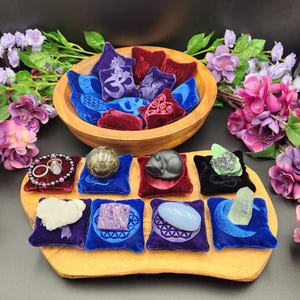 May include: A wooden bowl filled with small velvet pillows in shades of blue, purple, and red, each adorned with embroidered symbols. Below, a wooden tray displays various gemstones and jewelry on matching velvet pillows, showcasing a variety of colors and textures.