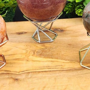 May include: Three spherical stones displayed on geometric metal stands. The left sphere is orange and white, the center is dark red, and the right is a dark grey sphere with red inclusions. The stands are rose gold, silver, and gold colored.