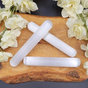 May include: Four white selenite wands are arranged on a wooden surface, with three wands crossed and one lying horizontally. White flowers are arranged around the wands. The wands have a smooth, cylindrical shape.