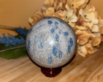 May include: A speckled gray sphere with blue markings, resting on a small, dark red wooden stand. The sphere is smooth and reflective, with a pattern of blue spots. Dried flowers and blue decorative elements are in the background.