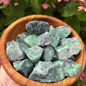 May include: A wooden bowl filled with rough-cut ruby in zoisite stones. The stones are a mix of green and black with flecks of red. The bowl is held in front of a background of green foliage and pink flowers.