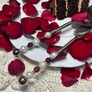May include: Cake server and knife set with pearl and gemstone handles. The handles feature alternating white pearls, brown and red gemstones, and silver accents. The blades are stainless steel. Red rose petals surround the set on a white plate.