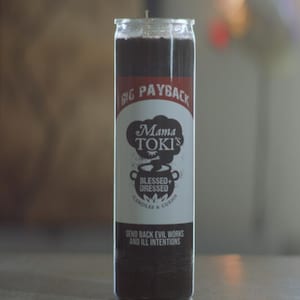May include: A black glass candle with a white label that reads "BIG PAYBACK Mama TOKI'S BLESSED+ DRESSED CANDLES & CURIOS SEND BACK EVIL WORKS AND ILL INTENTIONS".