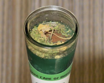May include: A green glass candle with a white label that says "Taking care of business" and "Mama Toki". The candle has a brown and green wax top with dried herbs and flowers.