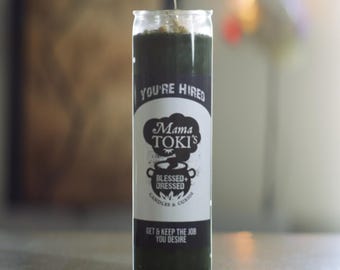 May include: A green glass candle with a white label that reads "You're Hired" and "Mama Toki's Blessed + Dressed Candles & Curios". The label also says "Get & Keep the Job You Desire".