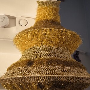 May include: A gold, crocheted Christmas tree ornament with a fluffy texture. The ornament is hanging from the ceiling.