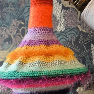 May include: A colorful crocheted lampshade with a wide, flared base and a narrow top. The lampshade is made of multiple stripes of different colors, including orange, purple, green, and pink. The lampshade has a fuzzy texture and is decorated with a fringe.