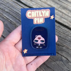 Pode incluir: Um alfinete Caitlyn em um cart&atilde;o azul. O alfinete apresenta uma ilustra&ccedil;&atilde;o estilizada de uma mulher com cabelo escuro, l&aacute;bios vermelhos e uma roupa preta. O cart&atilde;o tem o texto "CAITLYN PIN" em uma bolha rosa e um endere&ccedil;o web.