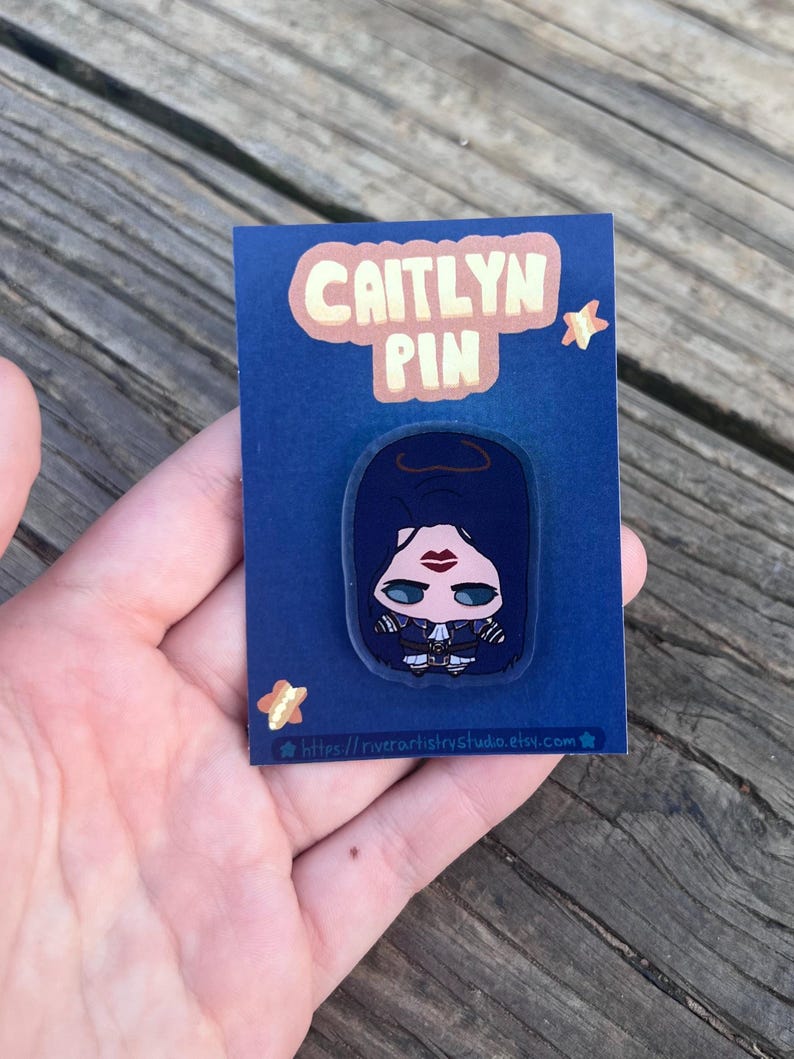 Pode incluir: Um alfinete Caitlyn em um cart&atilde;o azul. O alfinete apresenta uma ilustra&ccedil;&atilde;o estilizada de uma mulher com cabelo escuro, l&aacute;bios vermelhos e uma roupa preta. O cart&atilde;o tem o texto "CAITLYN PIN" em uma bolha rosa e um endere&ccedil;o web.