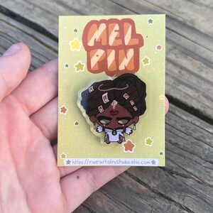 Pode incluir: Um alfinete de esmalte colorido com uma ilustra&ccedil;&atilde;o estilizada de uma pessoa com pele escura, vestindo uma roupa branca e joias de ouro. O alfinete est&aacute; em um cart&atilde;o amarelo com o texto "MEL PIN" e o URL da loja Etsy.