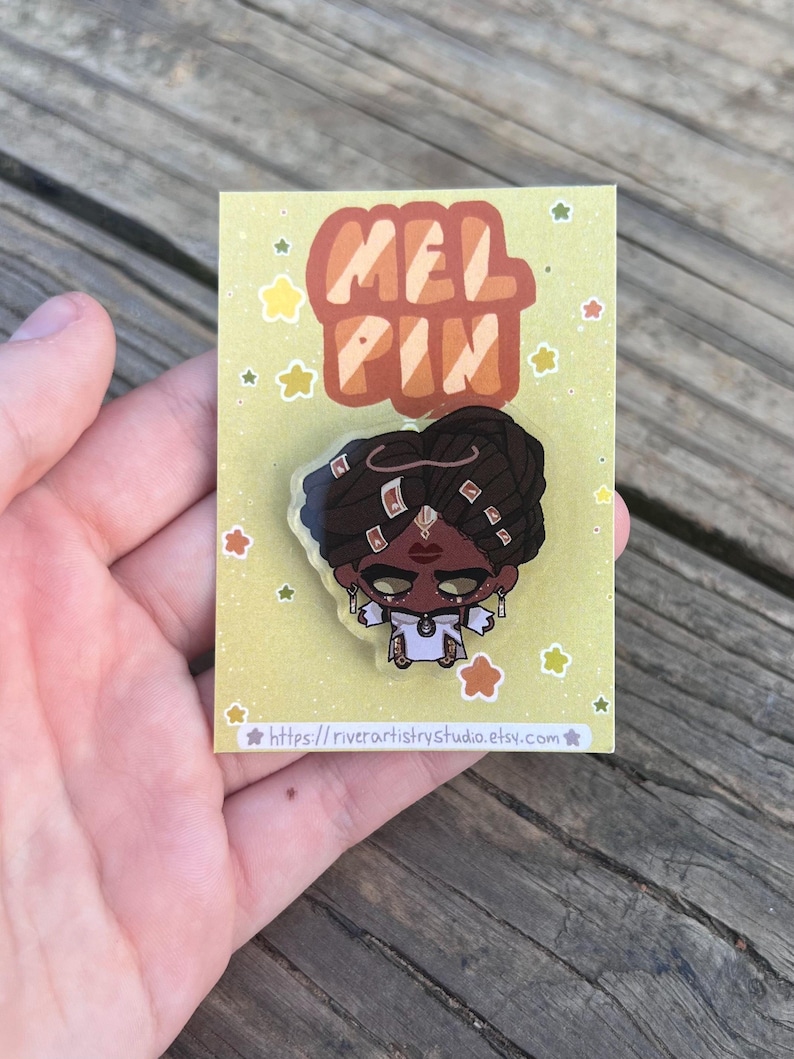 Pode incluir: Um alfinete de esmalte colorido com uma ilustra&ccedil;&atilde;o estilizada de uma pessoa com pele escura, vestindo uma roupa branca e joias de ouro. O alfinete est&aacute; em um cart&atilde;o amarelo com o texto "MEL PIN" e o URL da loja Etsy.