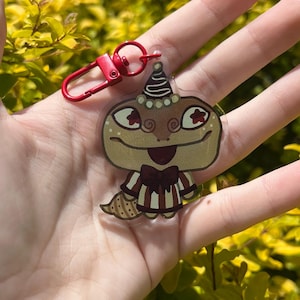 May include: A clear acrylic keychain featuring a cartoon lizard character with a party hat and bow tie. The lizard is brown, tan, and red, with a striped outfit. The keychain has a red clasp.