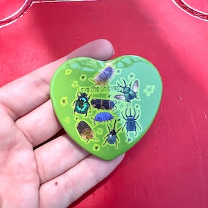 May include: A heart-shaped green pin featuring various colorful beetle and isopod illustrations. The text "Love the unloved" is printed in the center, surrounded by small yellow star graphics scattered across the smooth, glossy surface.