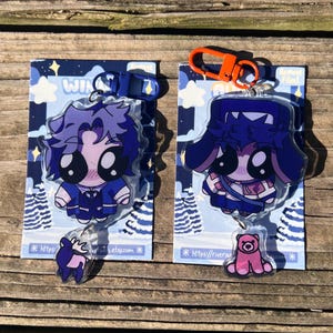 Op de afbeelding: Twee acryl sleutelhangers met cartoonfiguren in blauwe outfits. Eén heeft paars haar en een hertenbedel, de andere heeft een hoed en een roze berenbedel. Beide zitten op blauwe kaarten met een winterthema met het woord "WINTER".