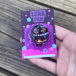 Pode incluir: Um alfinete de esmalte colorido com um personagem de desenho animado com a boca costurada e um chap&eacute;u. O alfinete est&aacute; em um cart&atilde;o de apoio roxo com o texto "ISHA PIN" e um endere&ccedil;o de site. O fundo &eacute; uma superf&iacute;cie de madeira.
