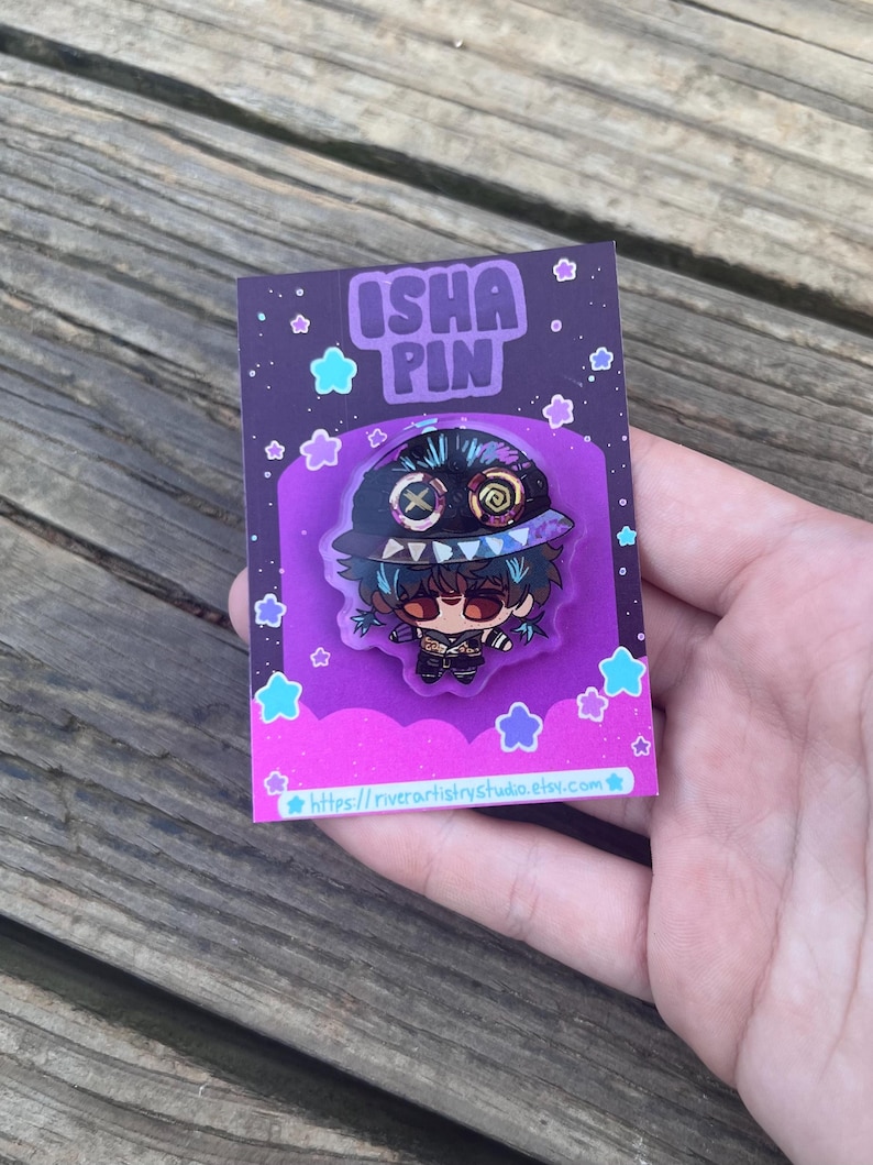 Pode incluir: Um alfinete de esmalte colorido com um personagem de desenho animado com a boca costurada e um chap&eacute;u. O alfinete est&aacute; em um cart&atilde;o de apoio roxo com o texto "ISHA PIN" e um endere&ccedil;o de site. O fundo &eacute; uma superf&iacute;cie de madeira.