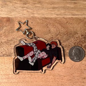 May include: A clear acrylic keychain featuring a cartoon illustration of two people relaxing on a red couch. The keychain has a gold star-shaped charm attached to a metal ring.
