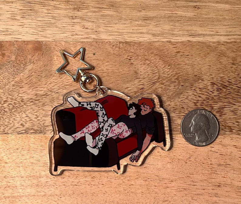 May include: A clear acrylic keychain featuring a cartoon illustration of two people relaxing on a red couch. The keychain has a gold star-shaped charm attached to a metal ring.