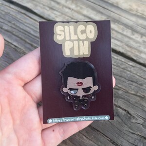 Pode incluir: Um alfinete Silco com uma ilustra&ccedil;&atilde;o em estilo cartoon de um personagem com cabelo preto, l&aacute;bios vermelhos e uma roupa preta. O alfinete est&aacute; em um fundo marrom com as palavras "SILCO PIN" em uma fonte bege.