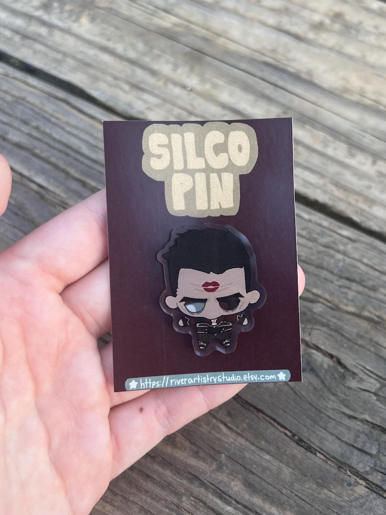 Pode incluir: Um alfinete Silco com uma ilustra&ccedil;&atilde;o em estilo cartoon de um personagem com cabelo preto, l&aacute;bios vermelhos e uma roupa preta. O alfinete est&aacute; em um fundo marrom com as palavras "SILCO PIN" em uma fonte bege.