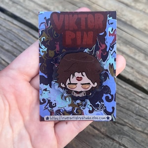 Pode incluir: Um alfinete com um personagem de desenho animado com cabelo castanho, batom vermelho e uma roupa preta. O alfinete est&aacute; em um cart&atilde;o com o texto "VIKTOR PIN" e um fundo azul e marrom rodopiante. O URL da loja Etsy &eacute; vis&iacute;vel na parte inferior.