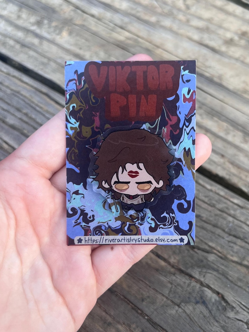 Pode incluir: Um alfinete com um personagem de desenho animado com cabelo castanho, batom vermelho e uma roupa preta. O alfinete est&aacute; em um cart&atilde;o com o texto "VIKTOR PIN" e um fundo azul e marrom rodopiante. O URL da loja Etsy &eacute; vis&iacute;vel na parte inferior.