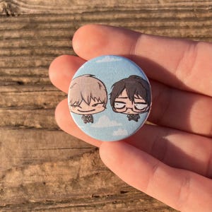 Può includere: Un bottone rotondo con sfondo azzurro chiaro con due personaggi dei cartoni animati con capelli chiari e scuri. I personaggi sorridono e fluttuano in un cielo con nuvole bianche. Il bottone è tenuto in una mano.