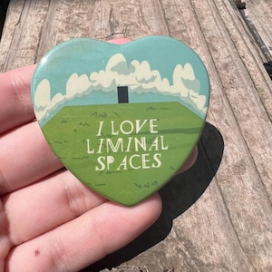 May include: A heart-shaped pin featuring a green grassy field, a blue sky with white clouds, and a small black rectangle in the center. The text "I LOVE LIMINAL SPACES" is printed in white capital letters across the lower portion of the pin.