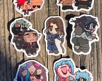 May include: A collection of colorful, pixelated stickers featuring various cartoon characters. The stickers are on a wooden surface and showcase a variety of designs, including characters with red, blue, and brown hair, and some with hats and accessories.