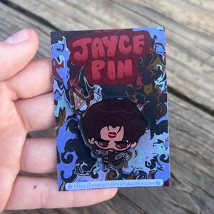 Pode incluir: Um alfinete com um personagem de desenho animado com cabelo castanho e um design de l&aacute;bios vermelhos. O alfinete est&aacute; em um cart&atilde;o com as palavras "JAYCE PIN" em letras vermelhas. O cart&atilde;o tem um design abstrato azul e marrom e o URL da loja Etsy.