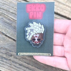 Pode incluir: Um alfinete com um personagem de desenho animado com cabelo branco, l&aacute;bios vermelhos e uma roupa vermelha e preta. O alfinete est&aacute; em um cart&atilde;o cinza com as palavras "EKKO PIN" em rosa. O URL da loja Etsy &eacute; vis&iacute;vel na parte inferior.