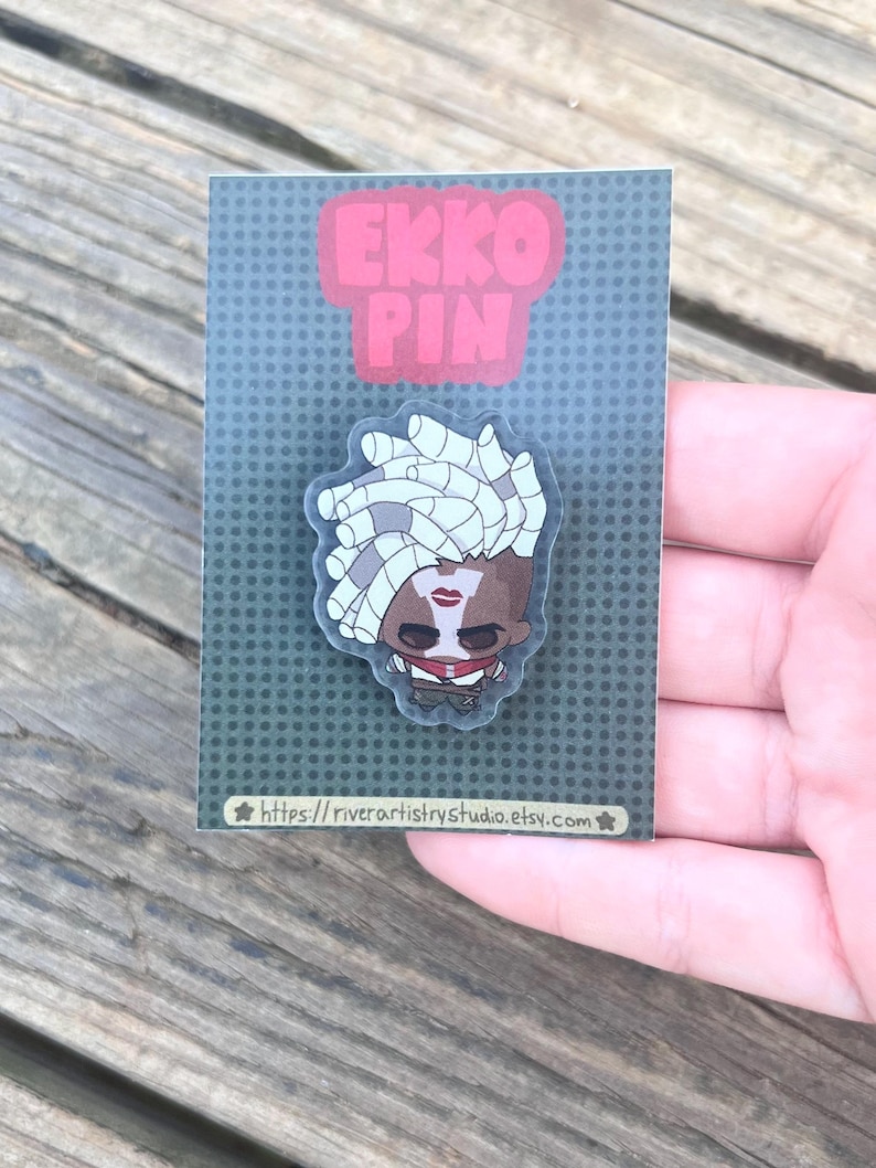 Pode incluir: Um alfinete com um personagem de desenho animado com cabelo branco, l&aacute;bios vermelhos e uma roupa vermelha e preta. O alfinete est&aacute; em um cart&atilde;o cinza com as palavras "EKKO PIN" em rosa. O URL da loja Etsy &eacute; vis&iacute;vel na parte inferior.