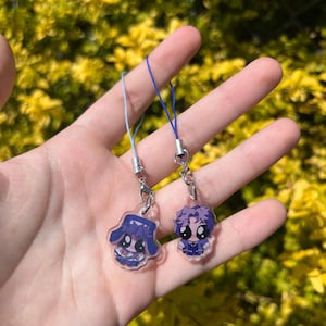 May include: Two anime-style charms with blue and purple designs. Each charm is attached to a silver-colored clasp and a blue or light blue string. The charms are held in front of a blurred background of yellow and green foliage.
