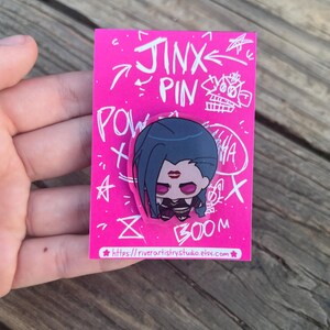 Pode incluir: Um alfinete rosa com um personagem de desenho animado com cabelo azul, batom vermelho e uma roupa preta. O alfinete est&aacute; em um cart&atilde;o rosa com as palavras "Jinx Pin" e outros rabiscos. O endere&ccedil;o do site &eacute; vis&iacute;vel na parte inferior.